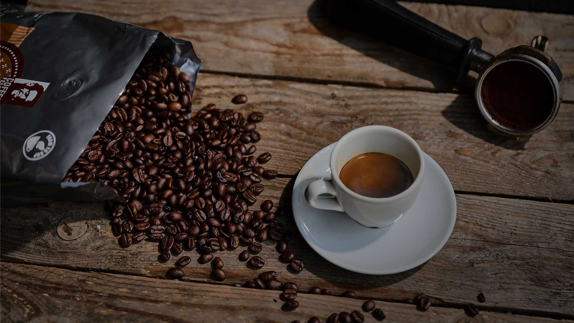 Geöffnete Tüte mit Kaffeebohnen, die auf dem Tisch liegt und Bohnen kommen raus, daneben steht ein Espresso 