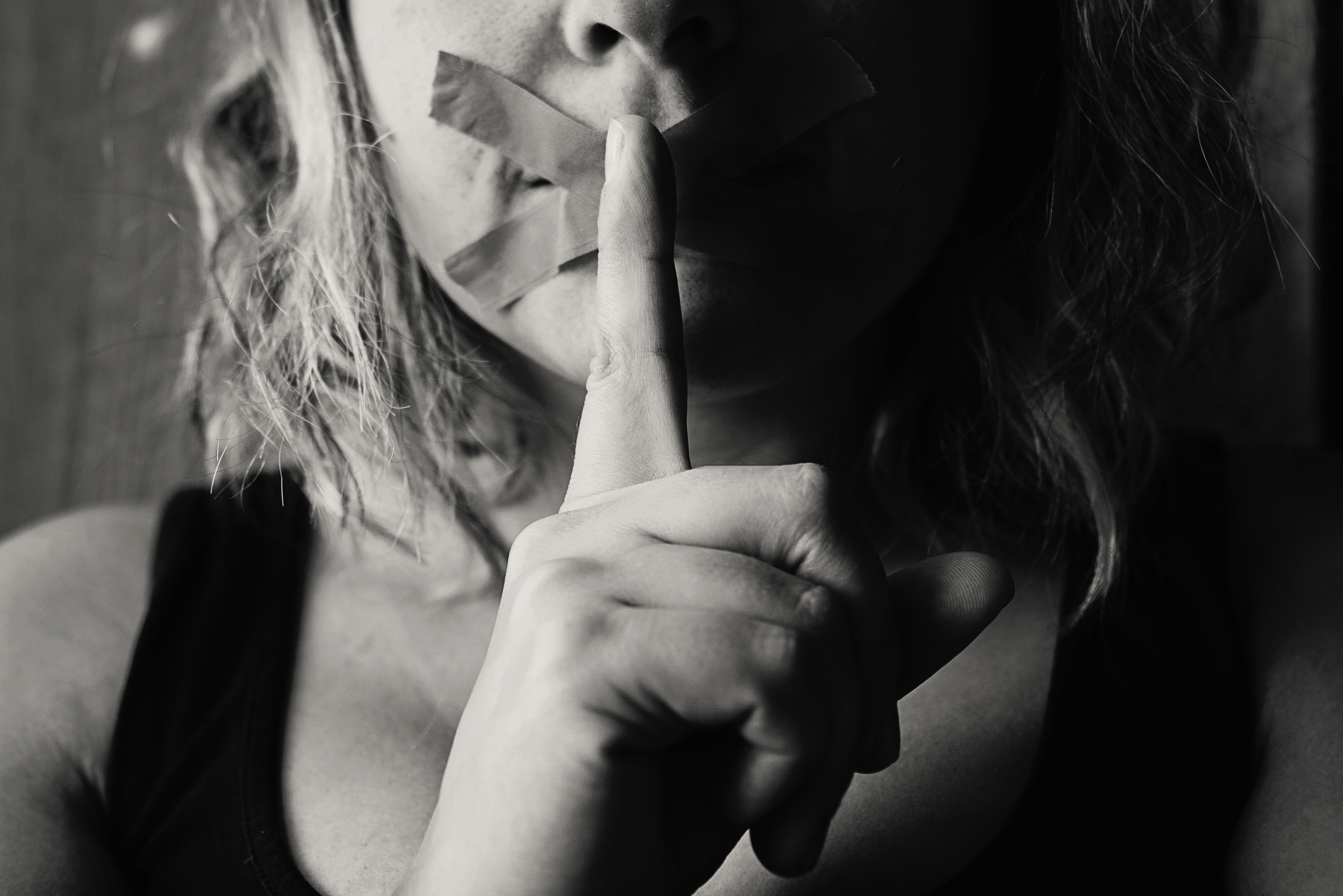 A black and white photo of a woman placing her finger over her lips. Her lips have been sealed by a plaster-like material