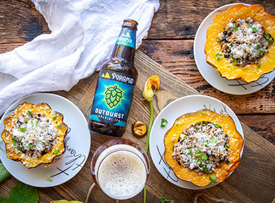 Two acorn squashes filled with sausage and kale on plates surrounded by a bottle of Outburst IPA