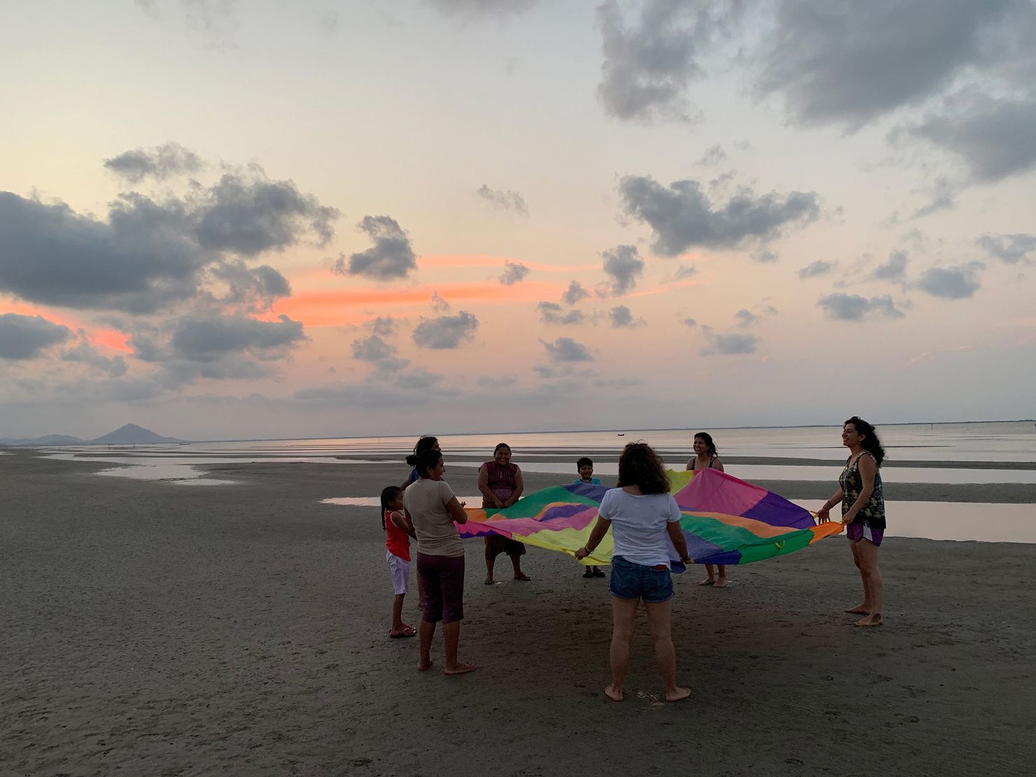 six women and two children stand on the beach at mar muerte holding a multicolored circular sheet
