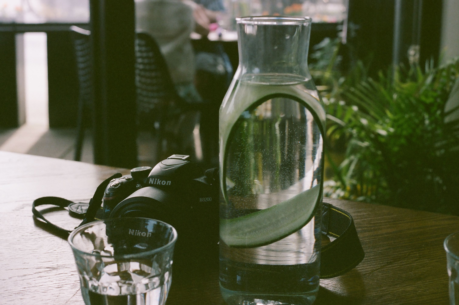 A jug of water with a long strip of cucumber in it, next to a digital camera.