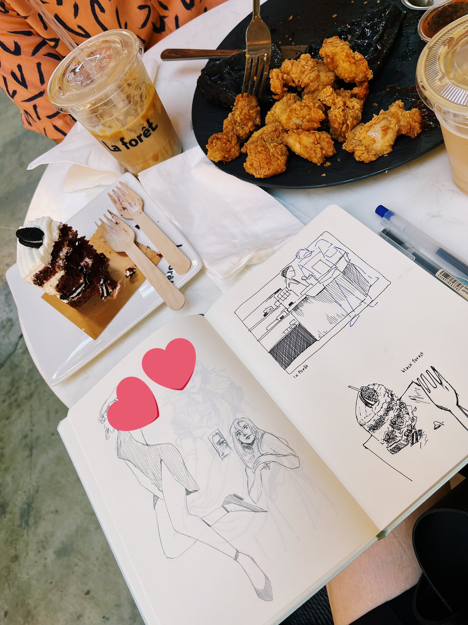 Sketchbook, cake, drinks, and fried chicken on a table. A heart emoji censors part of the sketchbook.