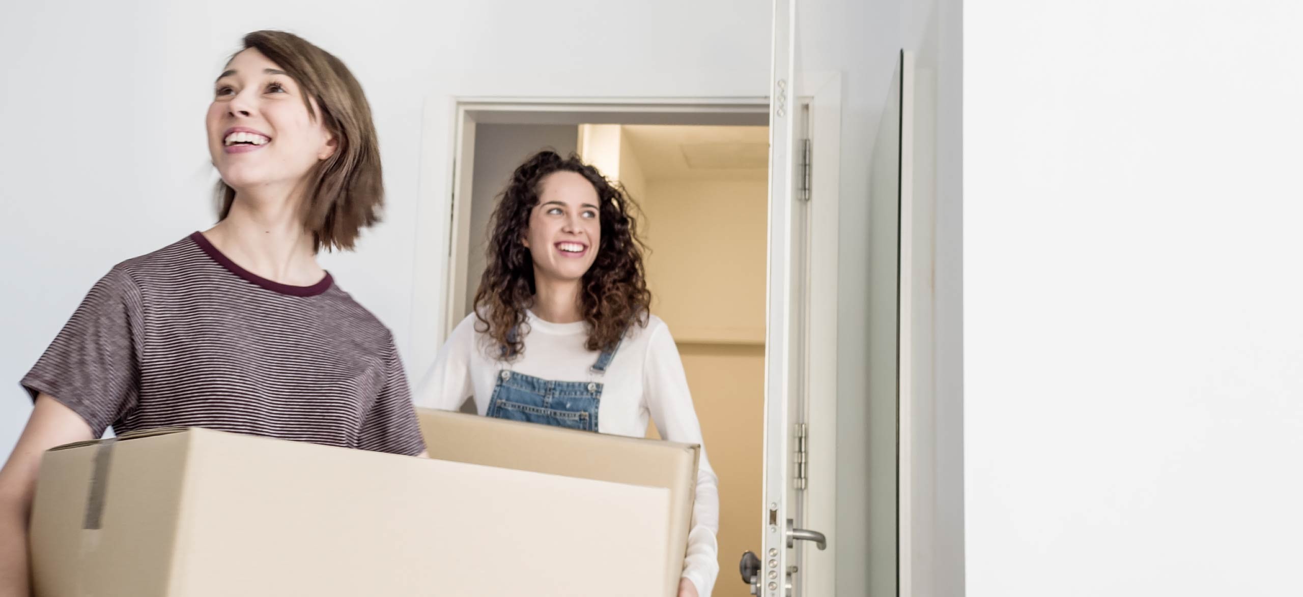 Two young people carry boxes into an apartment, smiling.