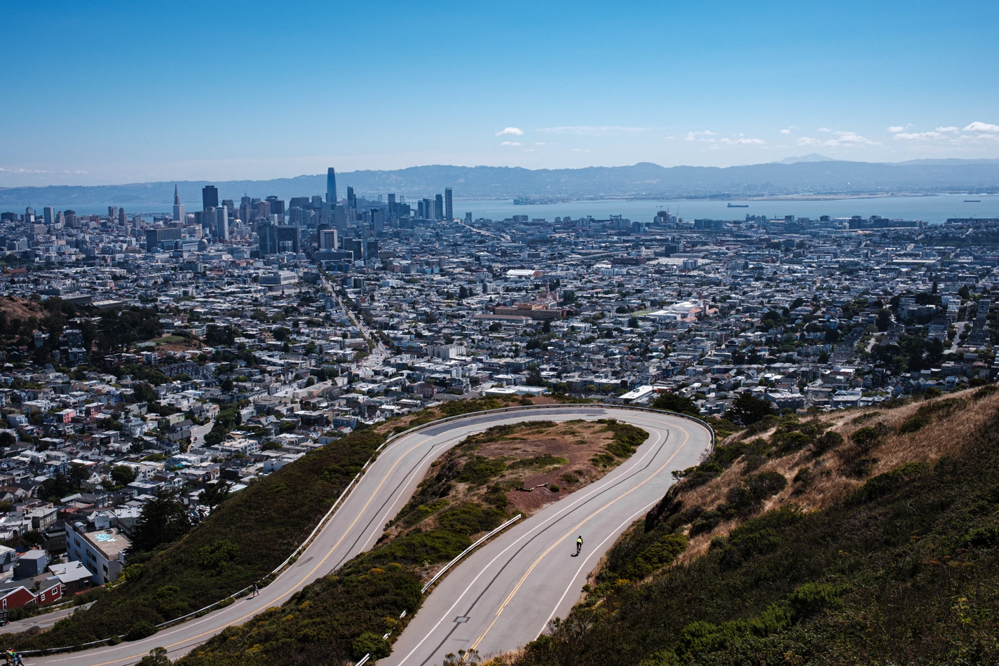 Downtown San Francisco, seen from Twin Peaks. A road curves around itself in the foreground and a lone cyclist can be seen as a tiny dot.
