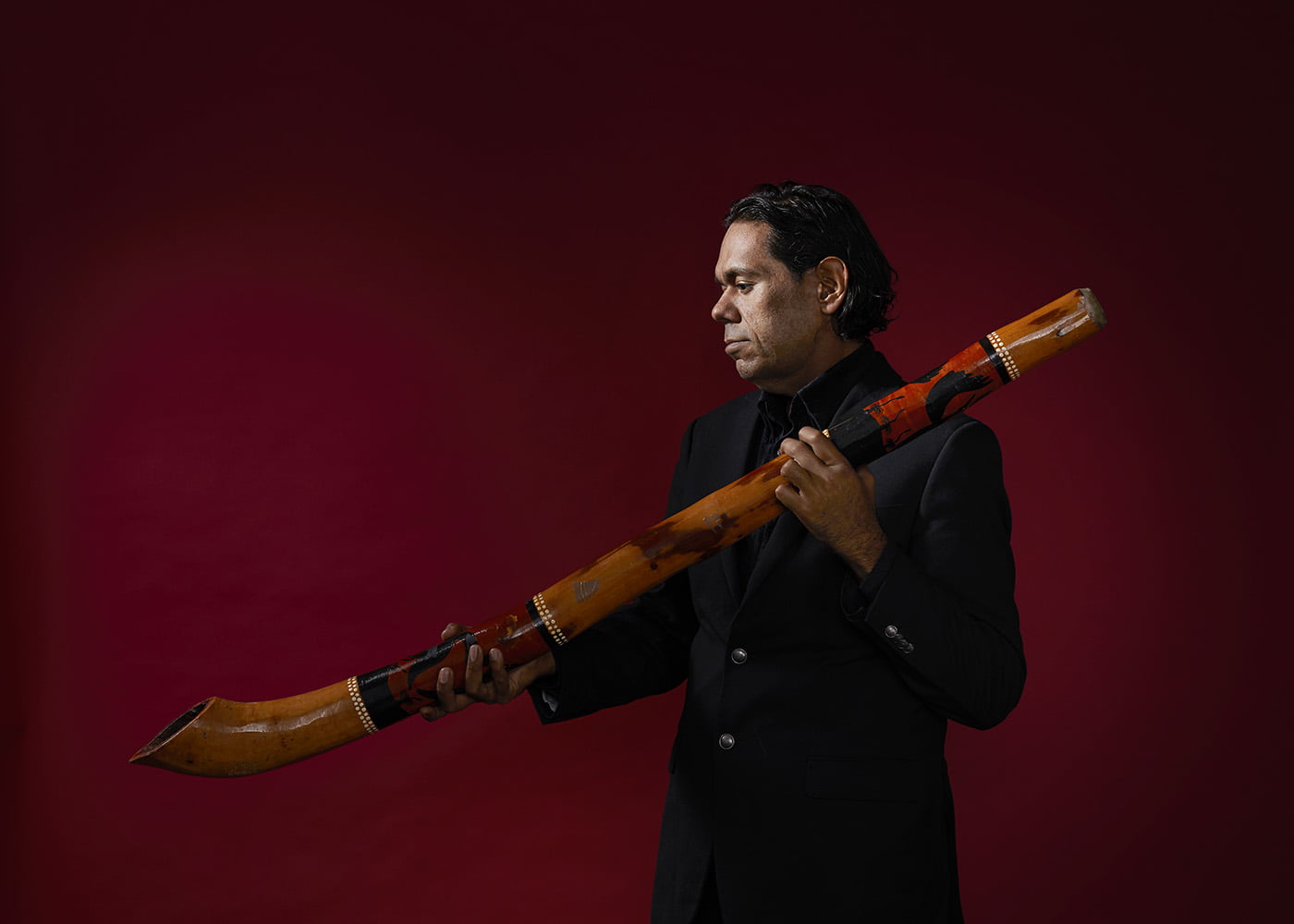 William Barton holding his didgeridoo in a studio in front of a burgundy background 