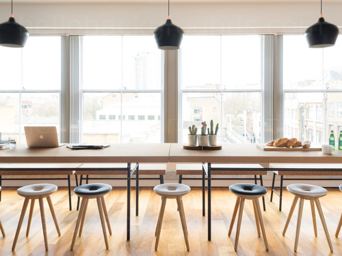 Contemporary wooden floored shared workspace containing wooden table and fabric topped stools next to large windows Skype background