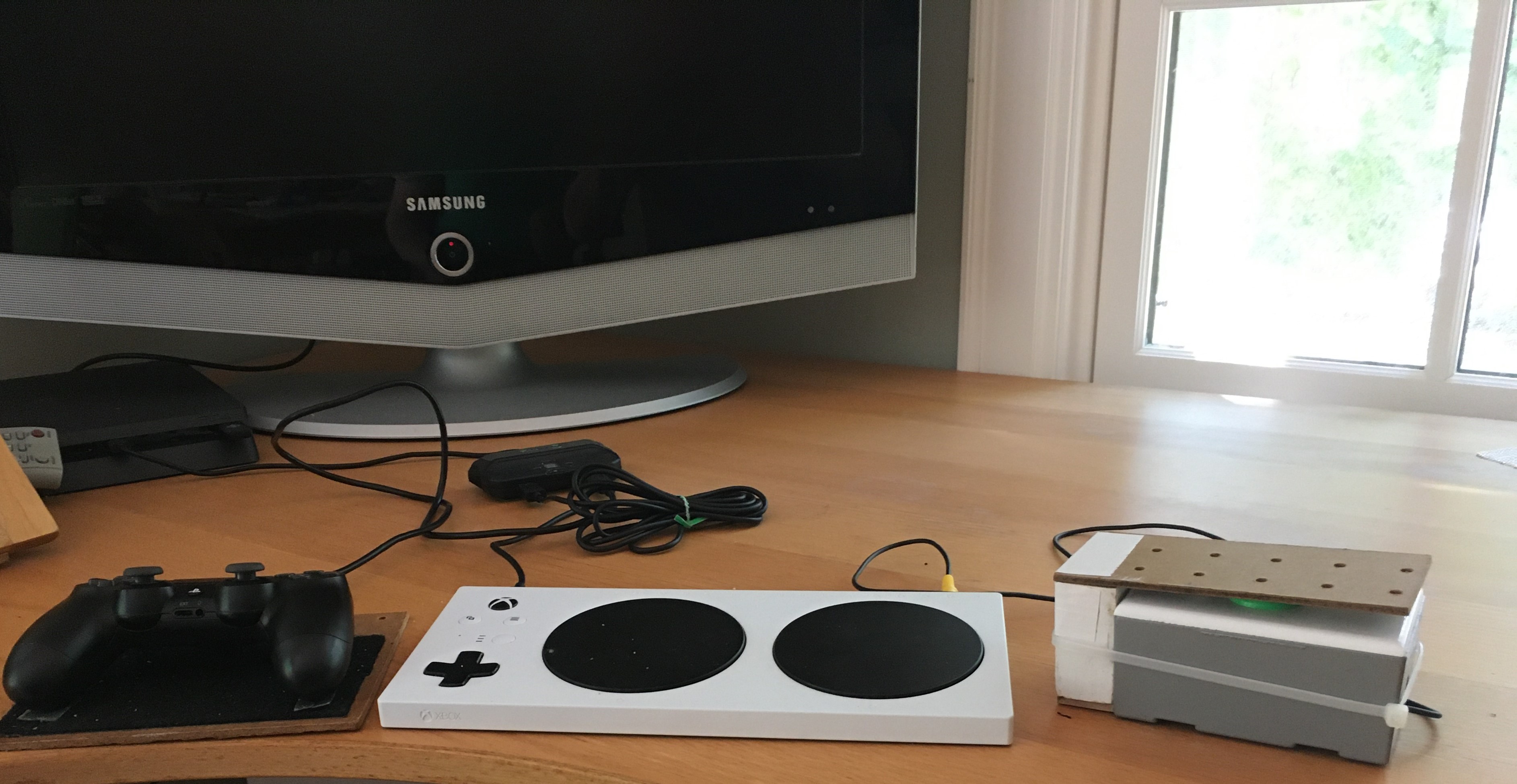 Sam Graves' Xbox Adaptive Controller laying on desk. To the left is a traditional video game controller attached to a wooden platform with Velcro on top. An external button switch is plugged in to the adaptive controller. Adaptive Controller and PlayStation console are plugged into the Titan Two adapter. A television is also on the desk.
