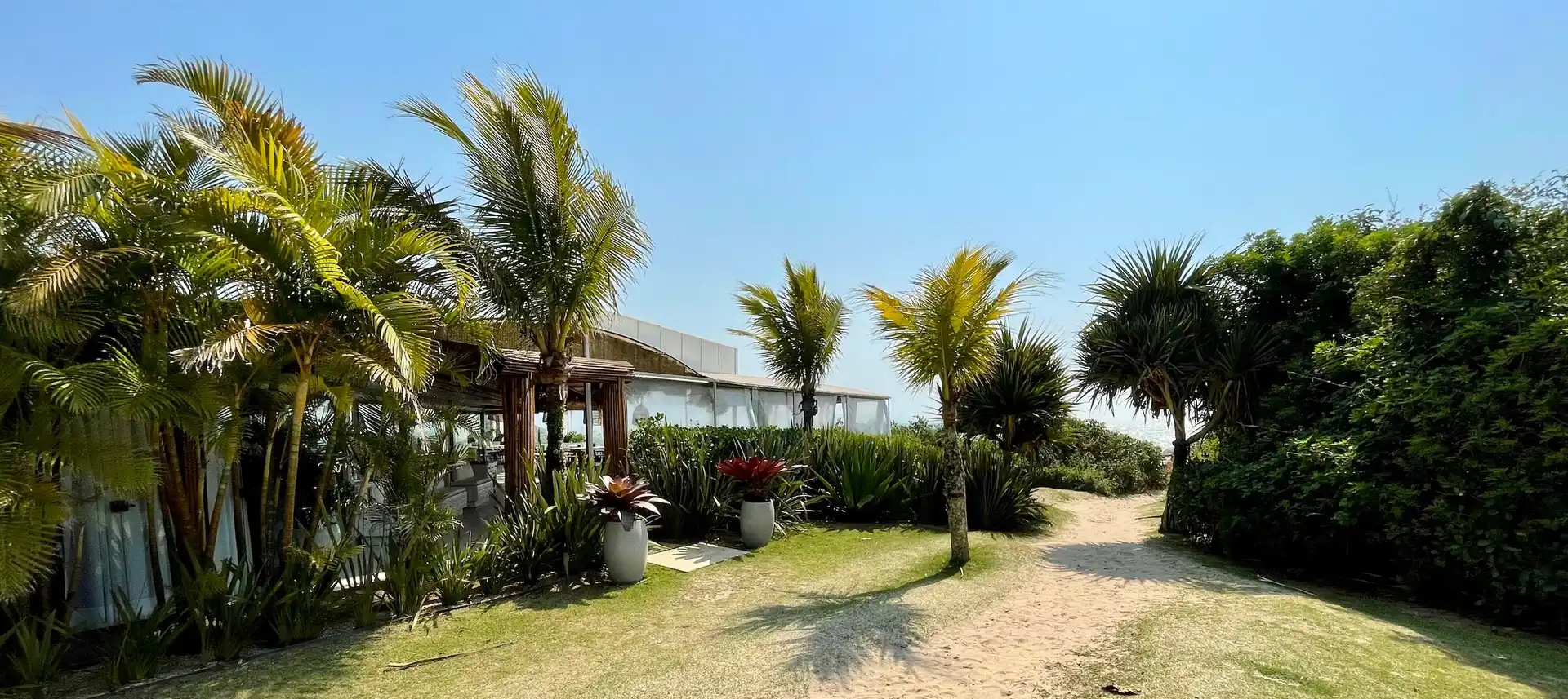 Vista do caminho de entrada na praia com coqueiros e restaurante a esquerda e céu azul