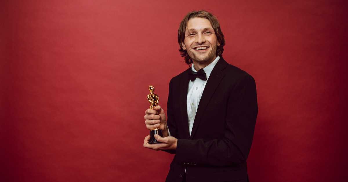 Ein Ehrenmitglied im Verein in einem vornehmen Anzug hält vor einer roten Wand eine Oscar-Figur in den Händen