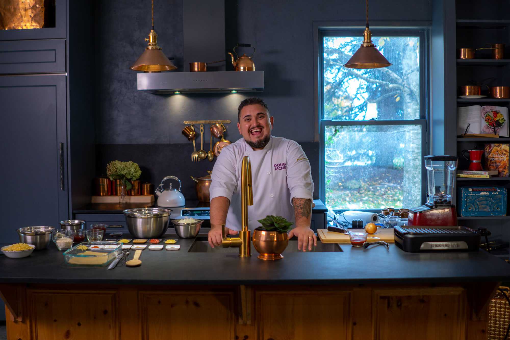 Chef Doug McNish laughs in a kitchen decorated with a black countertop and walls, with gold appliances.