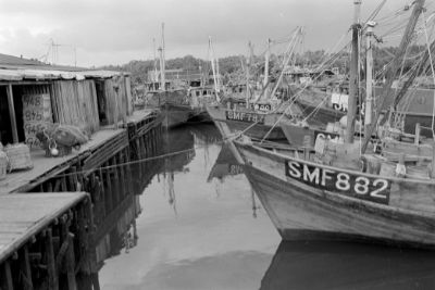 A black and white photo of fishing boats docked closely in a harbour in Kangkar. A row of sheds lie across from the boats, built on a wooden platform standing on stilts.