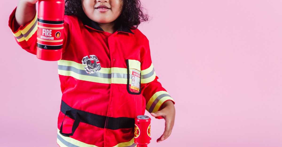 Ein Mädchen feiert Feuerwehr Geburtstag und ist als Feuerwehrfrau verkleidet, es hält einen Spielzeug-Feuerlöscher in die Luft