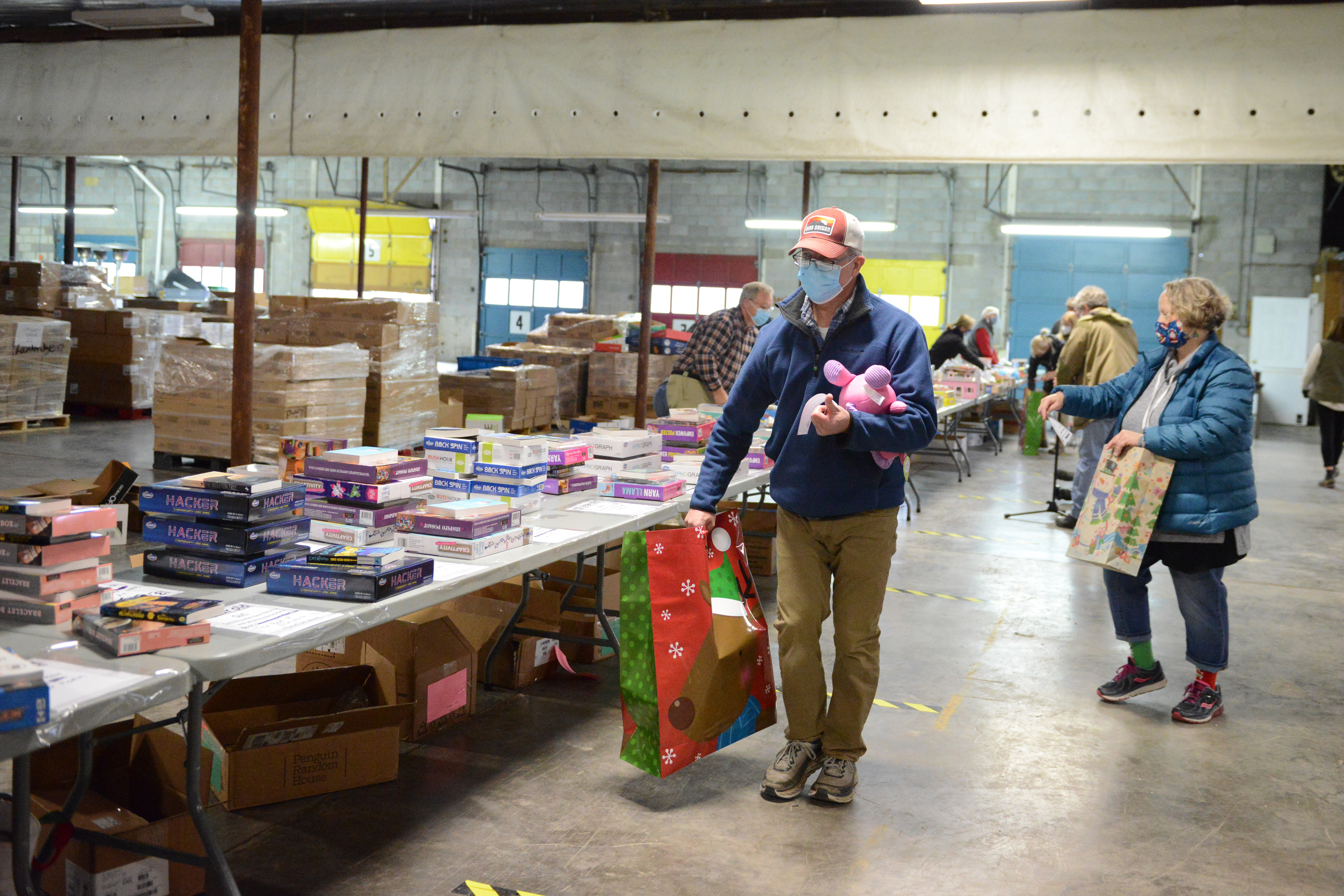 Adult male volunteer in the foreground, with pink stuffed animal in one arm, and a large Christmas gift bag with a reindeer in the other. Tracy Bonds, in the background, also packing toys into gift bags.