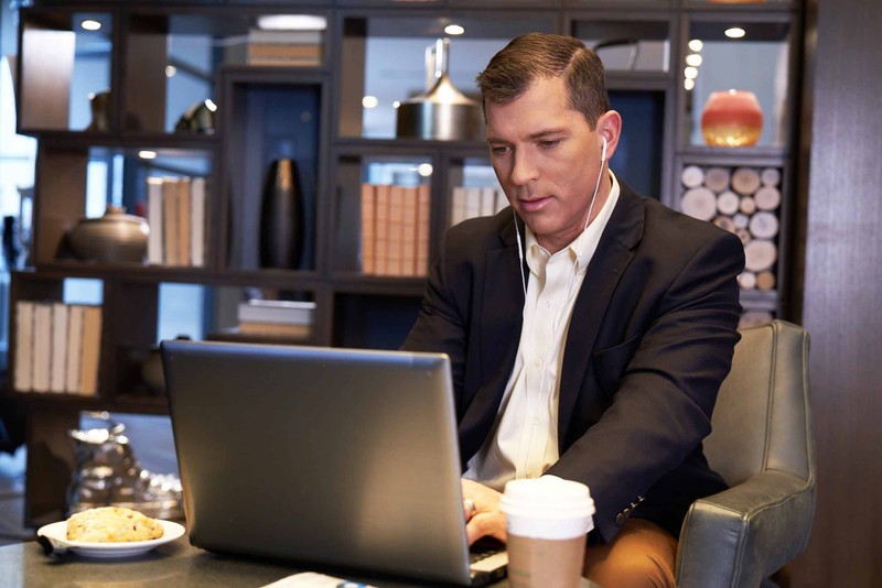 Man in a black blazer types on his laptop while listening to earbuds during a coffee break