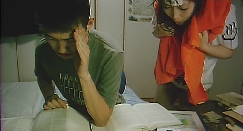 A screenshot from the film 'The Glamorous Life of Sachiko Hanai' of a boy at his desk doing his homework and hiding his face from a woman hovering above.