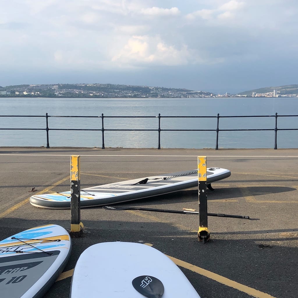 Three paddle boards on the ground, next to 2 black and yellow bollards. Black railings edge the path next to the sea. One the other side of the bay, there are hills and buildings.