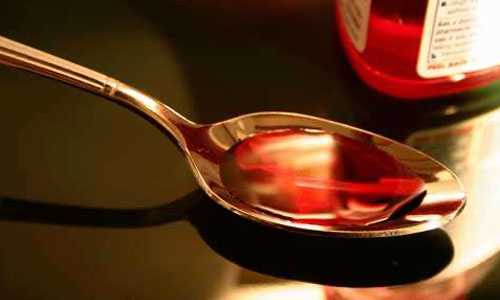 Photo of a bottle of cough medicine and a spoon filled with cough medicine on a dark countertop.