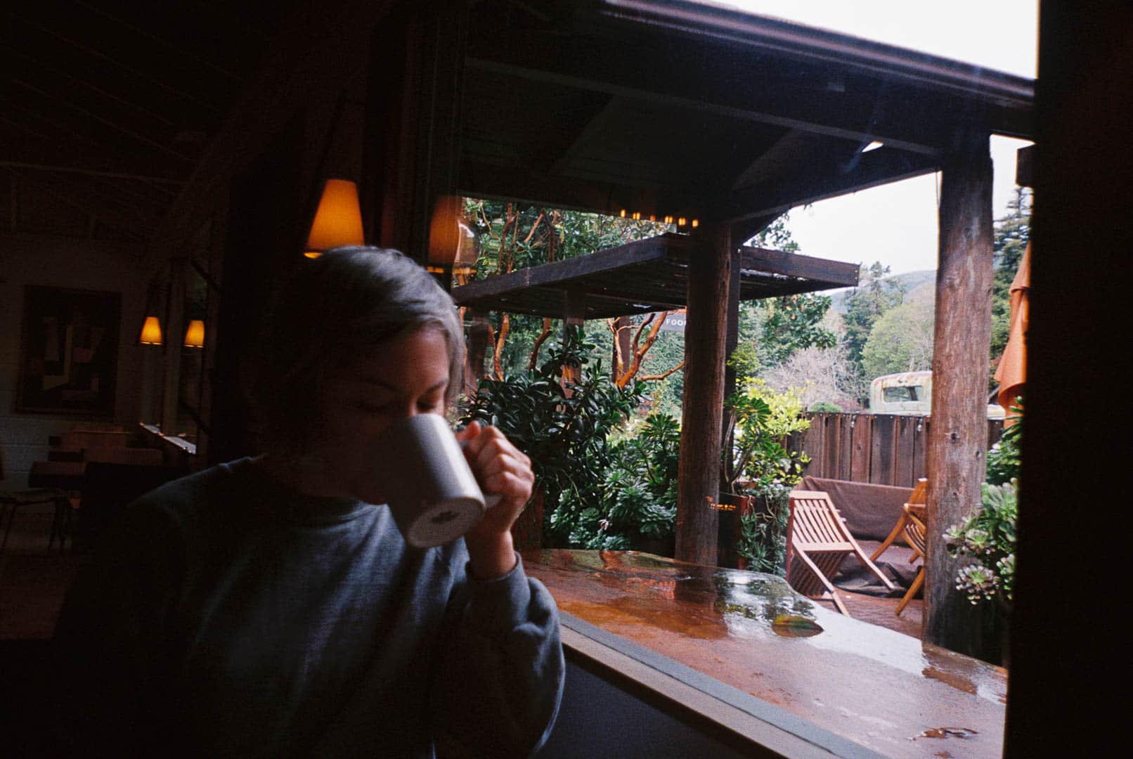 A woman sipping coffee next to a window overlooking a lush and damp garden area