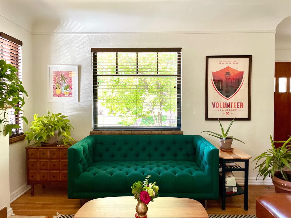 An emerald green sofa sits under a bright and sunny picture window. Art hangs on either side, and (surprise) plants sit on just about every available surface.
