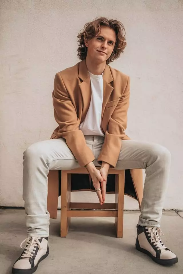 man in tan coat sitting on a chair with his hands clasped