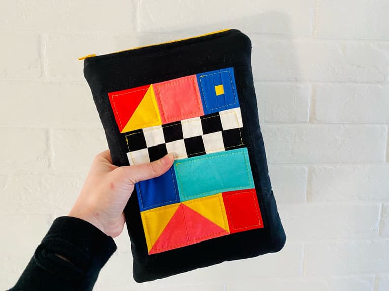 A black pouch with a patchwork panel made of triangles, squares, and rectangles. Colors include red, yellow, pink, blue, teal, black, and white. There are black and white squares in a checkboard pattern. Two red and yellow Posca markers are next to the pouch