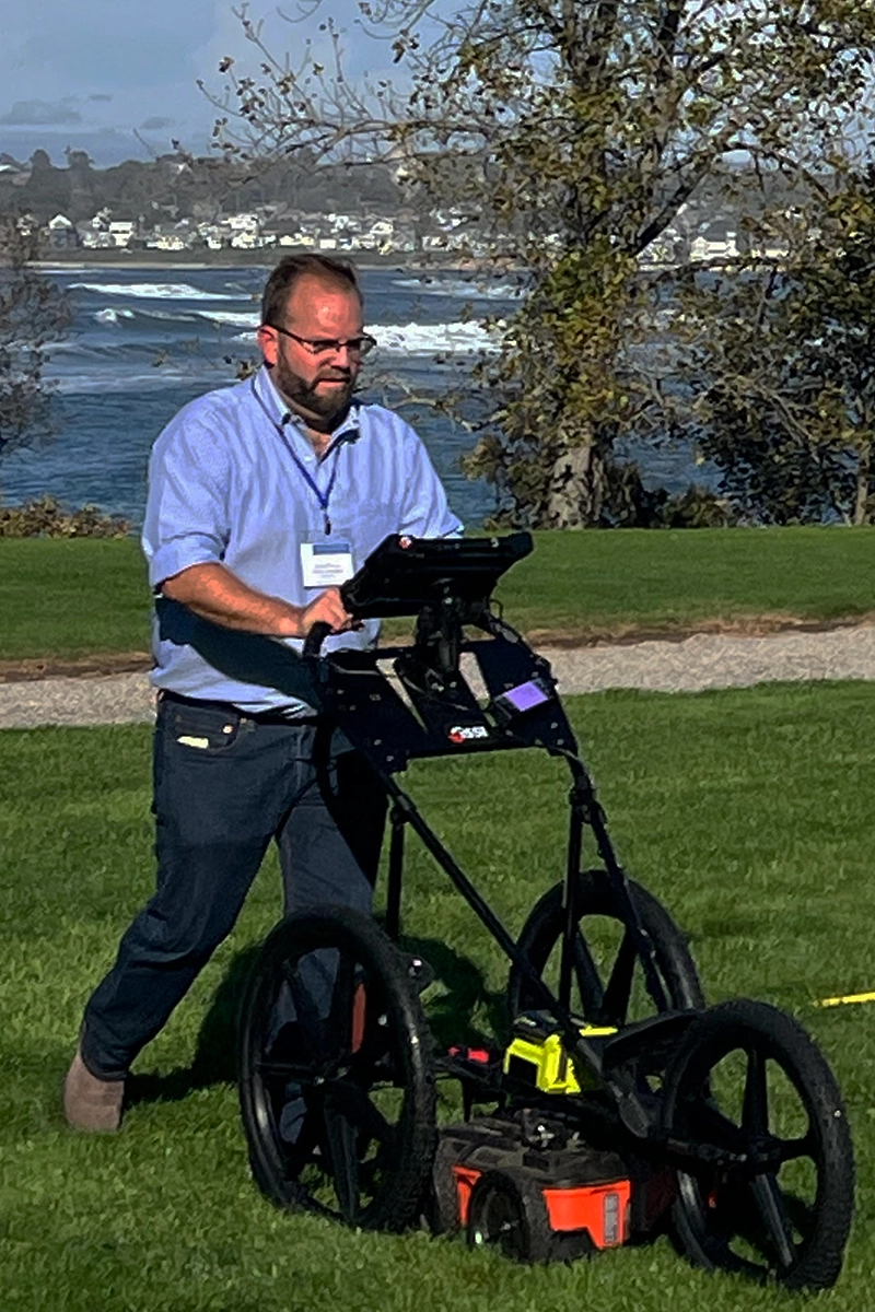 Man running a GPR machine over a field in front of the ocean.  