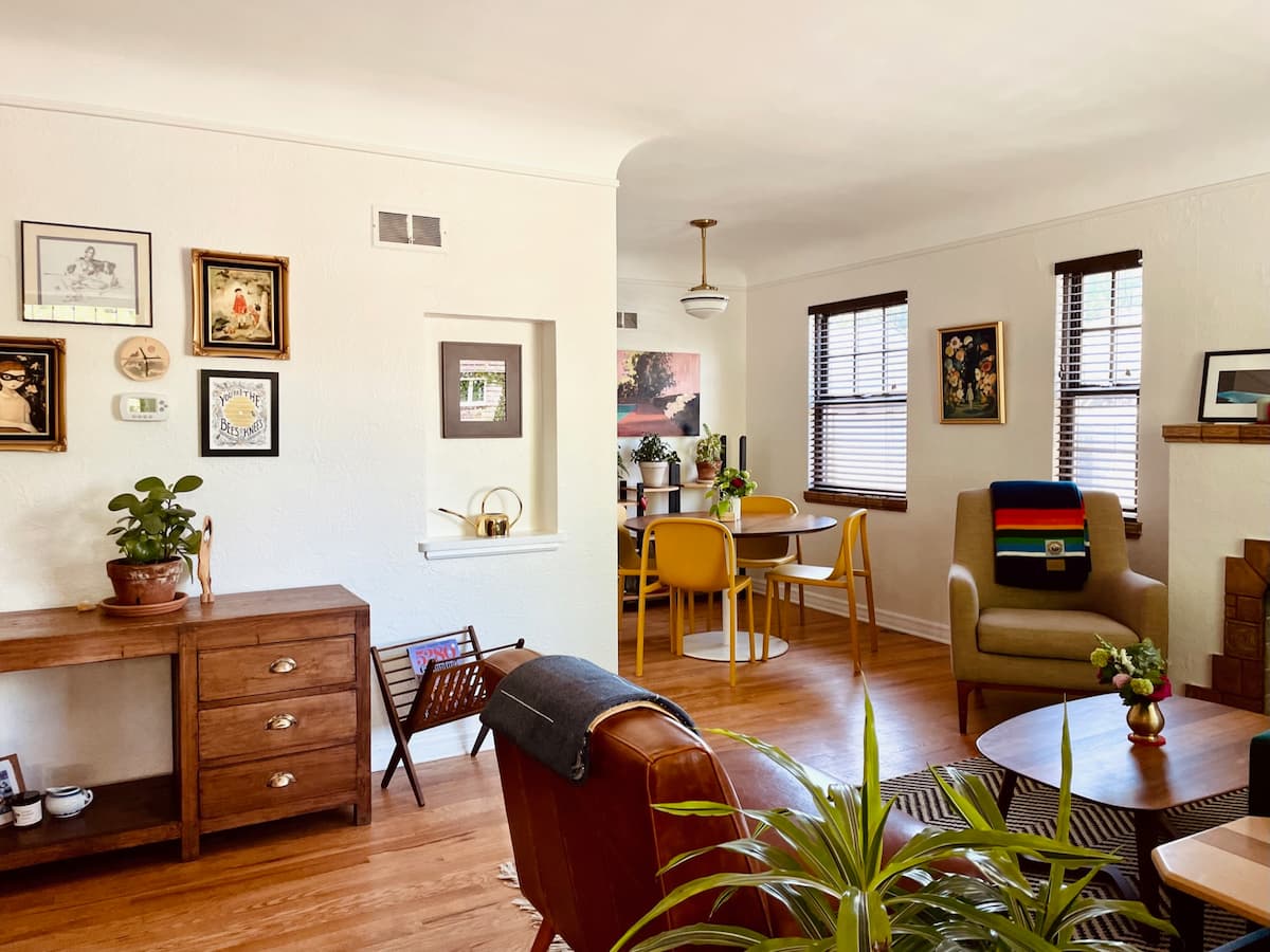 A warmly decorated living room with wood floors, peeking into the small dining nook. Art adorns the wall, plants sit on just about every available surface.