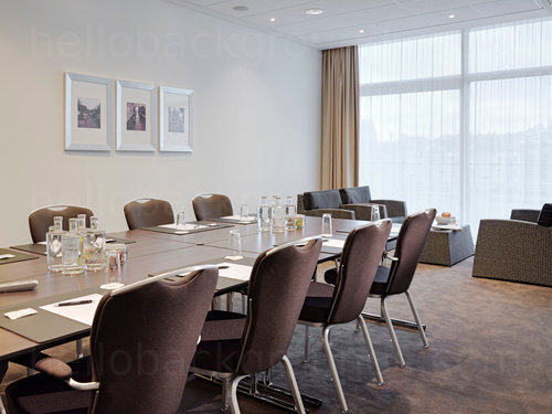 Conservative meeting room containing metal framed wooden table with brown fabric chairs and large floor to ceiling windows Microsoft Teams background
