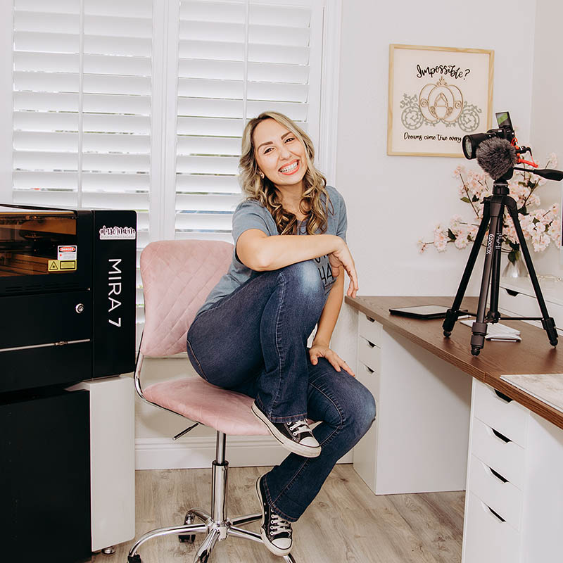 Emily sitting on an swivel chair smiling next to her Mira 7 laser, a desk with a camera and boom mic. On the wall behind her is a sign that reads, Impossible? Dreams come true everyday.