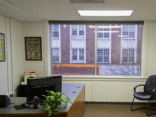Modern private office large window holding street views featuring corner style computer desk and black guests chair Zoom background