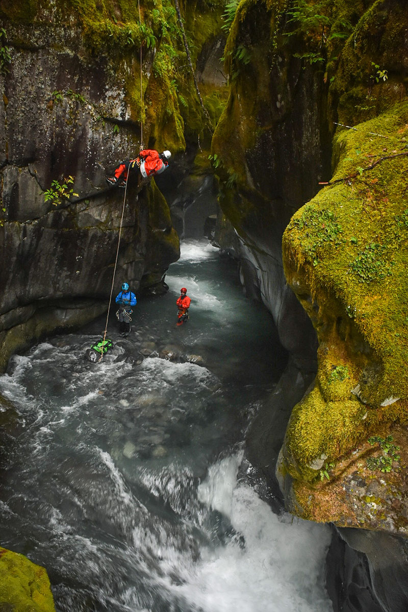 Zooming in, we see the man hanging from a rope in a canyon. Two other climbers are standing below him in a small creek at the bottom of the canyon