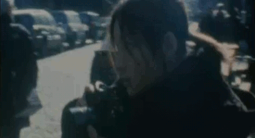 An animated gif of a scene from the movie 'Love Collage' of a grainy 8mm shot of a woman taking a photograph in the city.