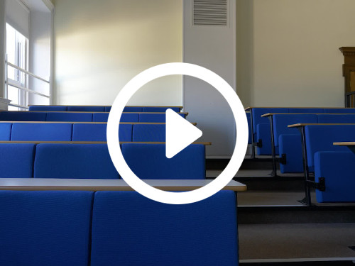 Simple lecture theatre style workspace with stepped rows of blue padded seating and white desks next to white framed window  Zoom background