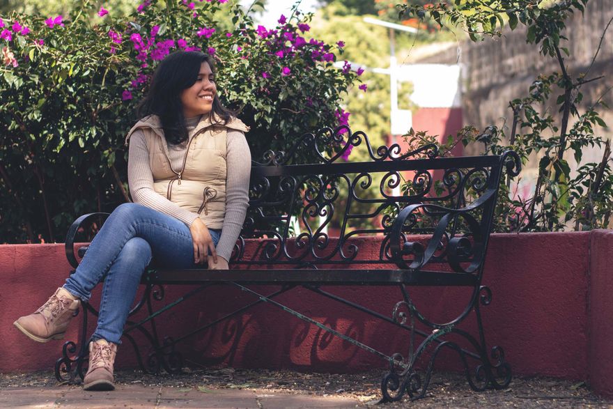 estrella, wearing jeans and a puffy vest, sits smiling on a black metal bench in front of a flowering bush