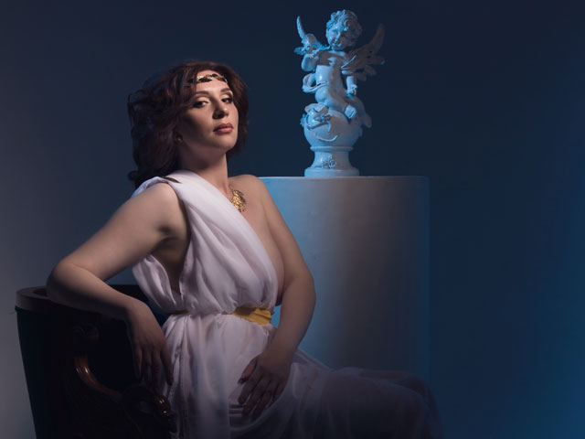 A Greek Goddess dressed in a Toga Costume sitting in front of an Ancient Statue of what appears to be Icarus