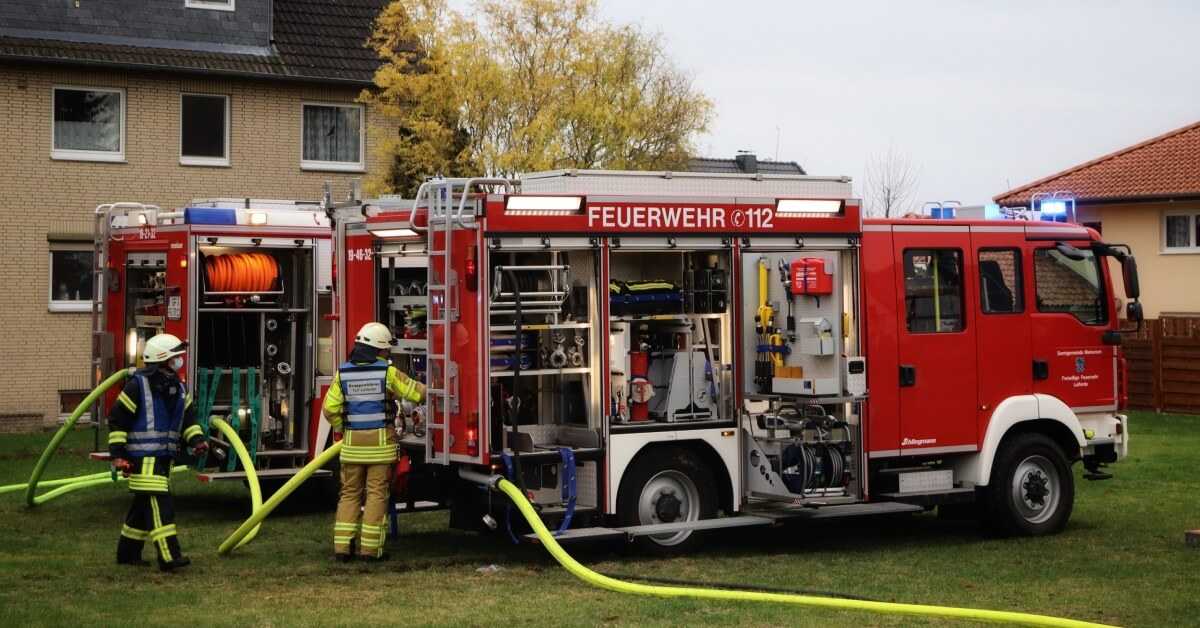 Zwei ehrenamtliche Feuerwehrleute bedienen einen Einsatzwagen, der vor einem Wohnhaus steht