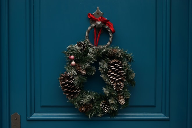 A holiday wreath made of evergreens and pinecones hangs from a round metal doorknocker attached to a teal door.