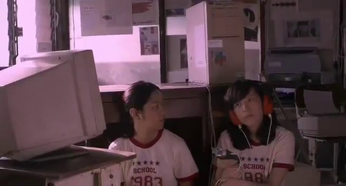 A screenshot of two girls in white and red school uniform t-shirts sitting on the floor next to a school computer. One of them is listening to headphones. From the movie 'Mary is Happy, Mary is Happy'.