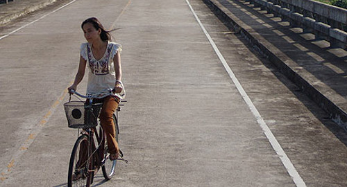 A screenshot from the film 'Wonderful Town' showing a woman riding her bicycle on the road alone.