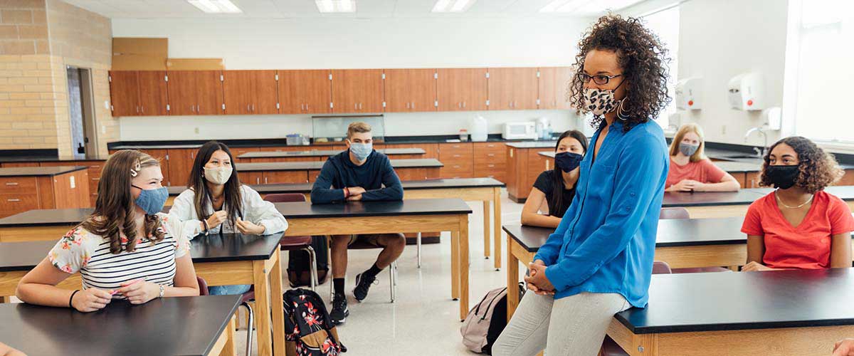 A teacher and students in a high school classroom, all wearing masks. Learn more about mandated reporter training requirements in California.