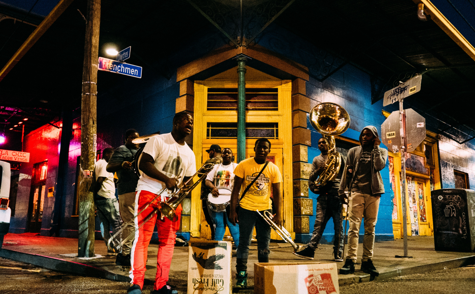 a group of musicians playing on a street corner in front of a blue and yellow building at night