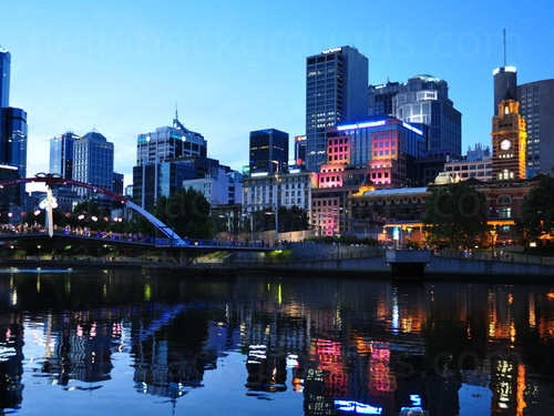 Harbourside urban cityscape at dusk featuring multiple buildings some lit by colourful lights next to modern style bridge  Skype background