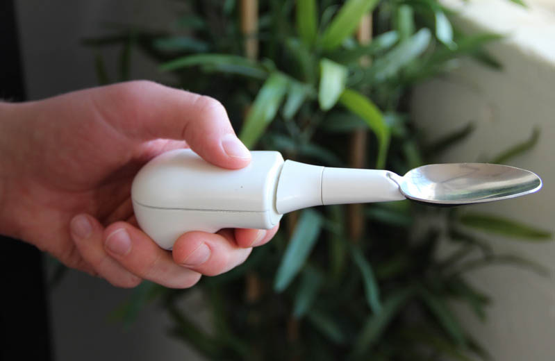 A closeup of a hand holding a white plastic-handles spoon, bifurcated in the middle to level the eating end, while the in-hand end receives and responds to even significant tremor shakes.