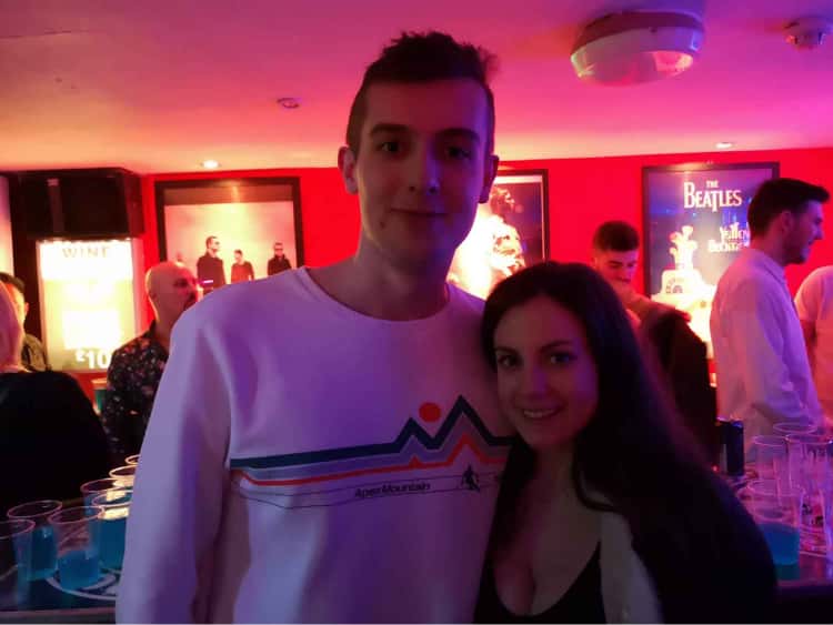 Myself in a white jumper next to my girlfriend, Naomi. We are stood in a bar in York called Stone Roses, with posters of various bands lining the walls behind us.