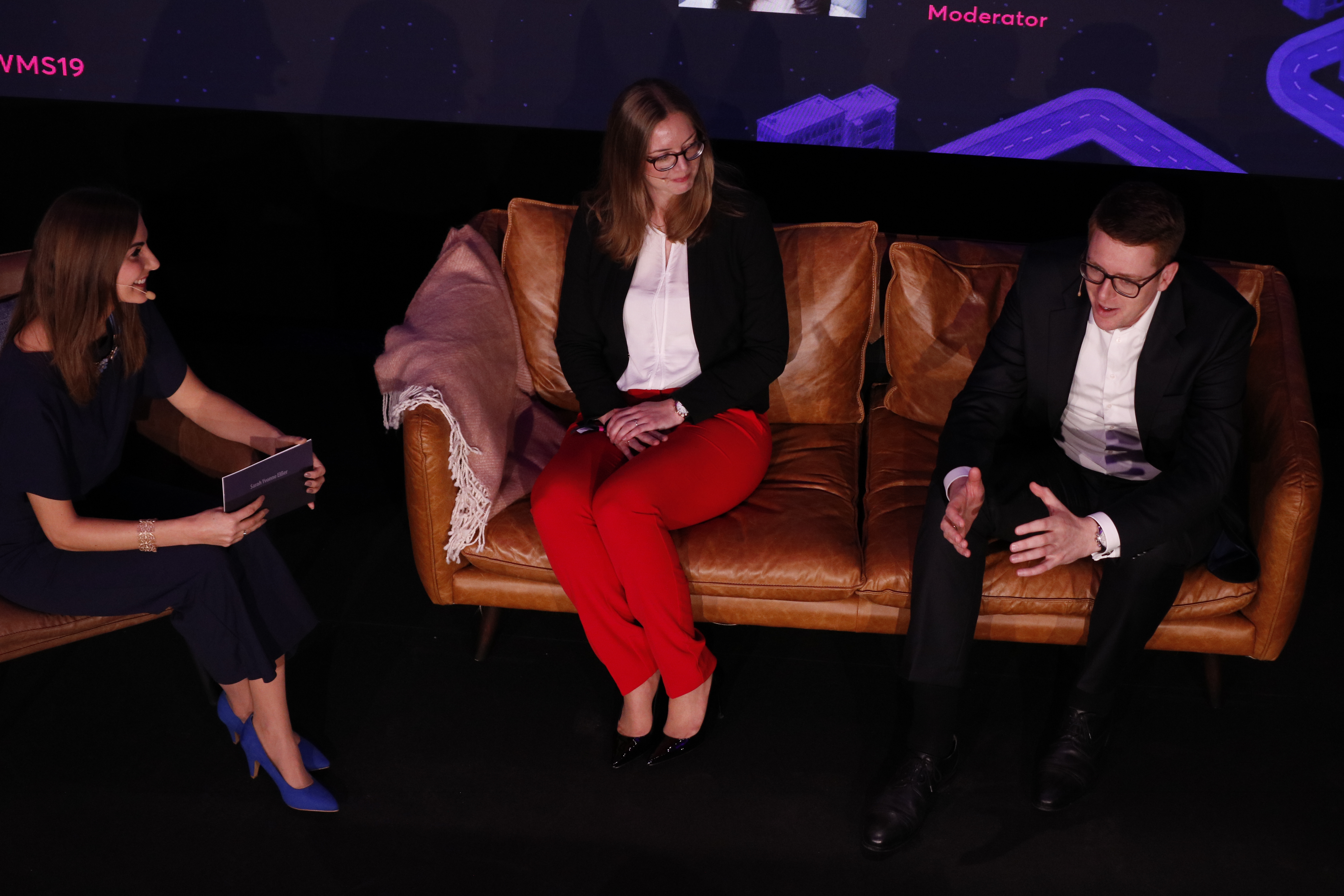 Two caucasian female persons and one caucasian male person sitting and speaking on a stage.