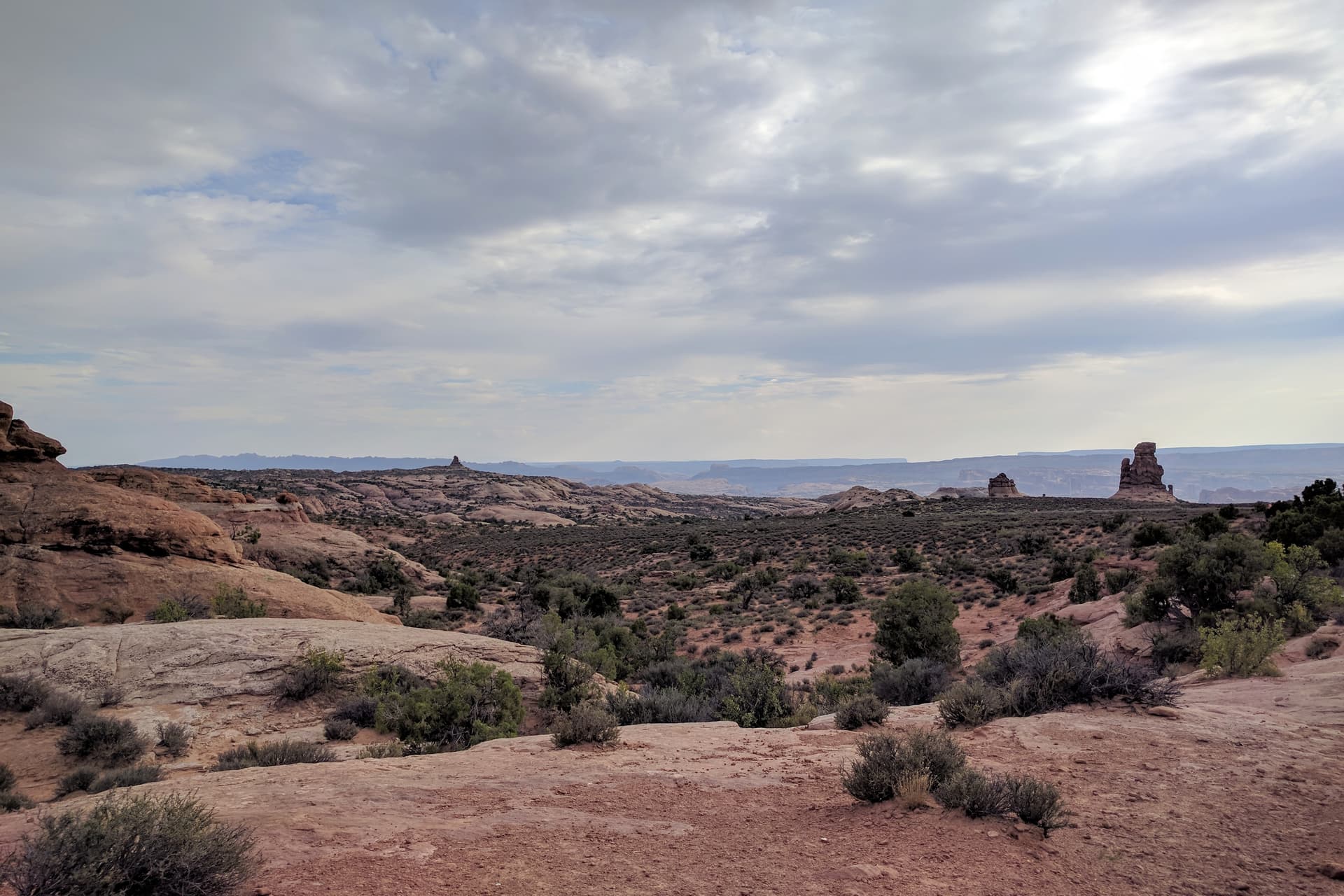 Looking east across Arches National Park. Red and white sandstone undulates across the top of the plateau, with a few stone pillars visible in the distance. On the horizon, the mesas and canyone of Canyonlands National Park.