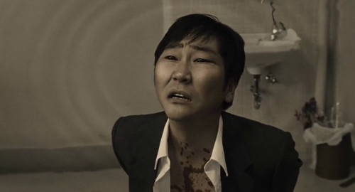 A screenshot from the movie 'R100' of a man in a roughed up suit with an alien expression on his face.