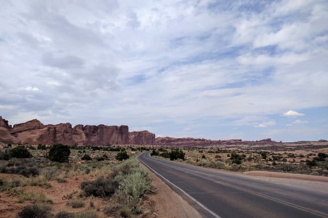 Looking north along the main road into Arches National Park. To the road's left, the undulating wall of a mesa made of red sandstone stretches into the distance.