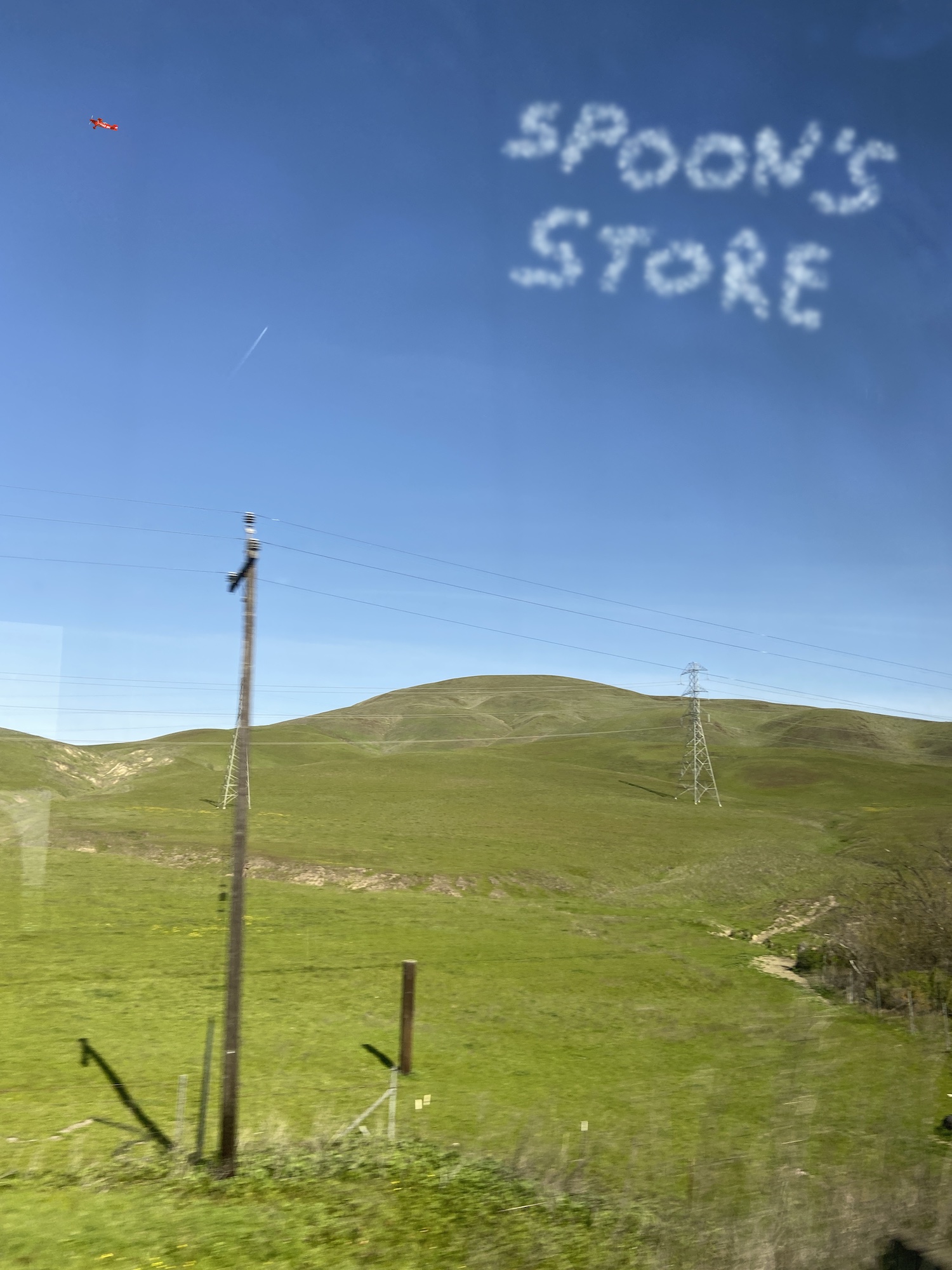 A green hill beneath a blue sky, with artificially superimposed skywriting letters that read Spoons Store.
