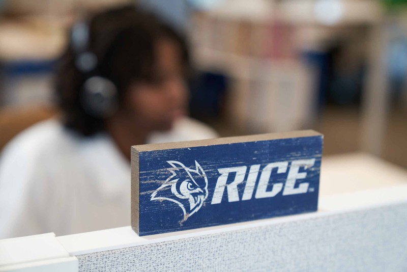Wooden block with Rice name and mascot on it set on top of a cubicle with student listening to headphones in the background